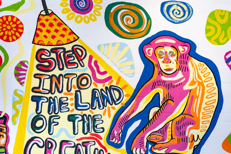 An illustration of a blue, yellow and pink monkey on a white wall, next to a red and yellow lamp shining a light on 'Step into the land of the' written underneath. The next word in the sentence is cut off from the photo.